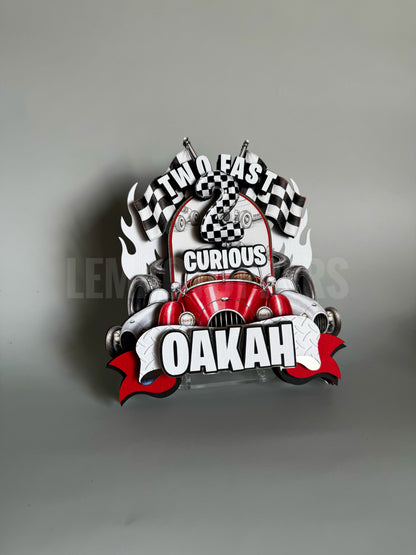A decorative metal sign featuring a stylized red race car with black-and-white checkered flags, flames, and bold text reading 'TWO FAST CURIOUS OAKAH' on a gray background.